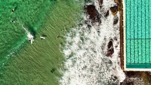 Aerial shot looking down onto ocean pool as person swims and surfers surfing in the crystal clear ocean in Bondi Beach, Sydney Australia. Birds Eye view.  Water crashes on rocks.