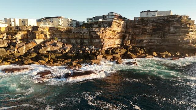 Aerial rolling view of houses perched on top of the dramatic seaside cliffs in North Bondi Sydney Australia.  Ocean crashes onto rocks below. 