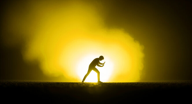 Silhouette of a person pushing against a bright yellow light. Man straining against an invisible force in a smoky, glowing environment. Conceptual image of perseverance and overcoming obstacles.