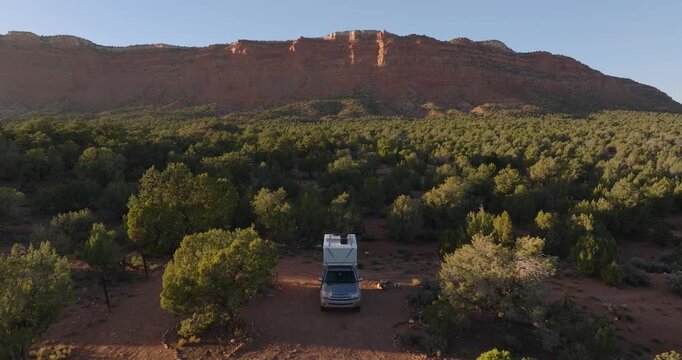 Camper van parked among green desert juniper trees beneath red sandstone mesa near Kanab Utah, drone dolly at sunset