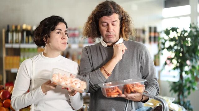 Couple choosing compare varieties cooled fresh shrimps, doubt and think before buying. Client choose and decide purchase prawns package, inspect good. Shop visitor pick up food in minimarket