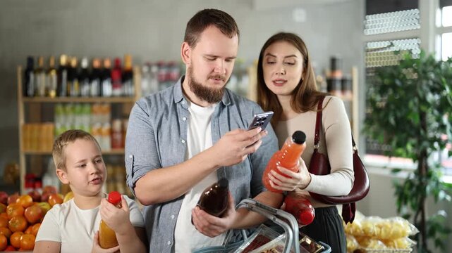 Friendly young parents with preteen son making purchases in local grocery store, using smartphone to scan barcodes on bottles of juices. Modern shopping and family teamwork concept