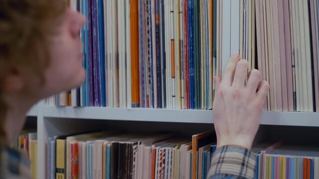 A close-up of a hand moving along a shelf of colorful books, looking for a needed resource. The library setting suggests research, study, or leisurely reading.