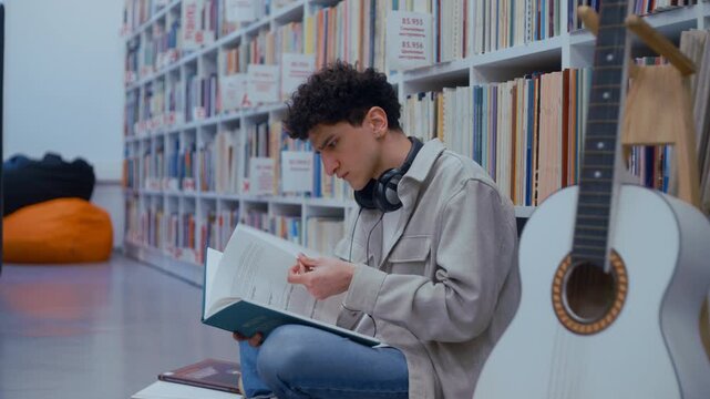 Young man sitting on the floor of a library, intently reading a music score from a book
