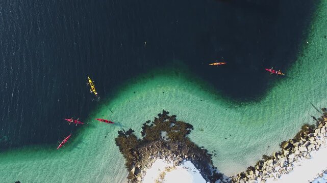 Top down drone view of people kayaking in turquoise arctic waters near a snowy fishing harbor, Lofoten Islands, Norway. Winter adventure and outdoor sports in scandinavian wilderness