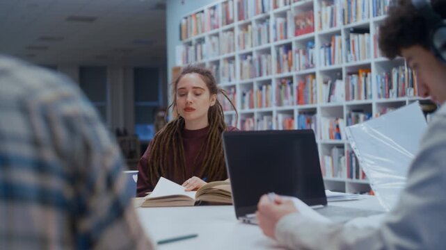 Young female student reading a book while studying with classmates in a university library