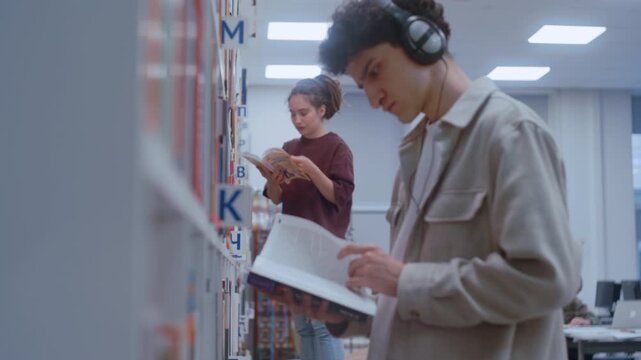 Two college students search for study materials among the tall wooden bookshelves of their campus library. 