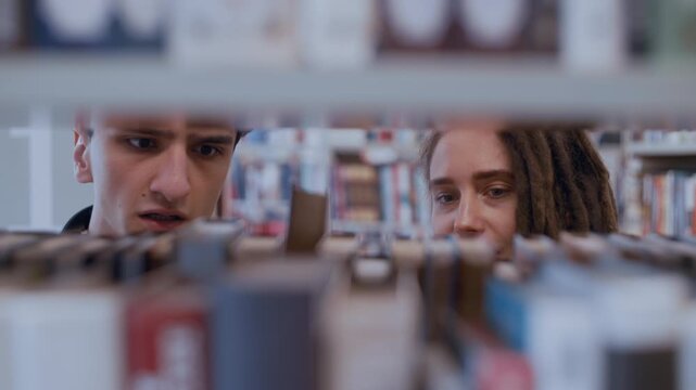 Young couple of college students looking for books on a bookshelf in a public library