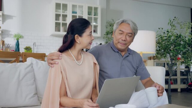 Senior spouses using laptop together sitting on couch at home. Elderly couple, hug and love on sofa with happy marriage, support and conversation of retirement home