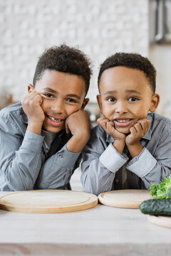 Two young brothers with curly hair rest their chins on their hands, smiling warmly at the camera in a bright kitchen setting with cutting boards and vegetables