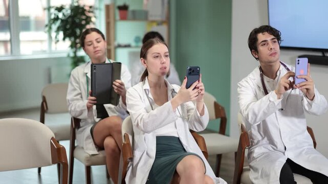 Focused group of medicals students sitting on chairs and listening lecture, taking photo or QR with mobil or tablet in light lecture hall