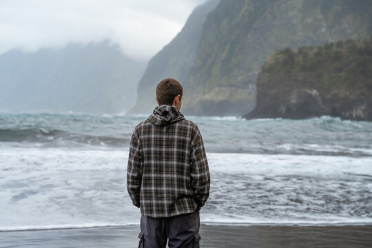 A man wearing a plaid hooded shirt stands on a dark sand beach, looking out at the crashing ocean waves and misty, steep mountains in the background under a cloudy sky.