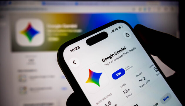 A person holding a smartphone displaying the google gemini app page on the apple app store, with a blurred computer screen in the background
