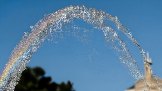 parabolic. A fountain's water jet arching against a blue sky with sunlight creating glints. travel magazines, destination branding, designed for travel destination branding, drives exploration.