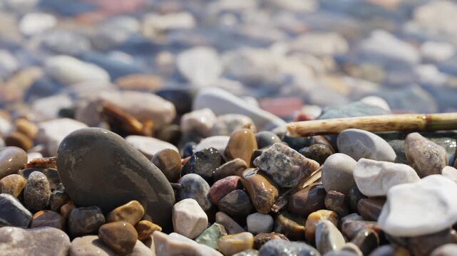 Close-up of colorful pebbles on a beach with crystal clear water gently washing over them in slow motion, creating a serene and relaxing natural background with sparkling light