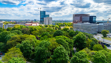 Sunny aerial drone view of Poznan cityscape with lush green trees of Marcinkowski Park in the foreground, modern skyscrapers, office buildings, construction crane, and cloudy blue sky. © Adam