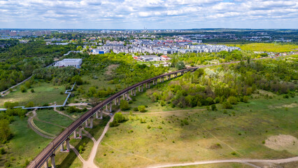 Fototapeta premium Aerial drone view of a long concrete railway viaduct in Poznan stretching across green fields and trees, with a modern city residential district and urban landscape in the background under a cloudy sk