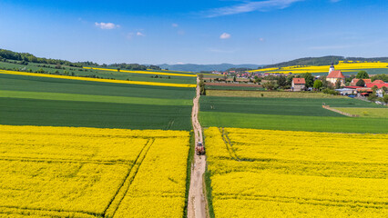 Wide aerial drone view of a farm tractor driving on a dirt road that divides vibrant yellow blooming rapeseed fields and green crops, with a village, church, and hills in the distance. © Adam