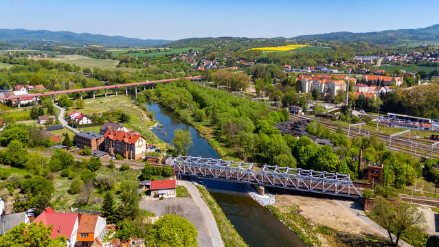 Wide aerial drone view of a metal truss railway bridge and a high road viaduct crossing over the Nysa Klodzka river in Klodzko, surrounded by residential buildings, train tracks, lush greenery, and hi