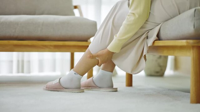 Female adult experiencing leg pain, muscle cramp, or discomfort in her calf. Woman sitting at home, touching her aching leg, seeking relief from physical strain or fatigue.