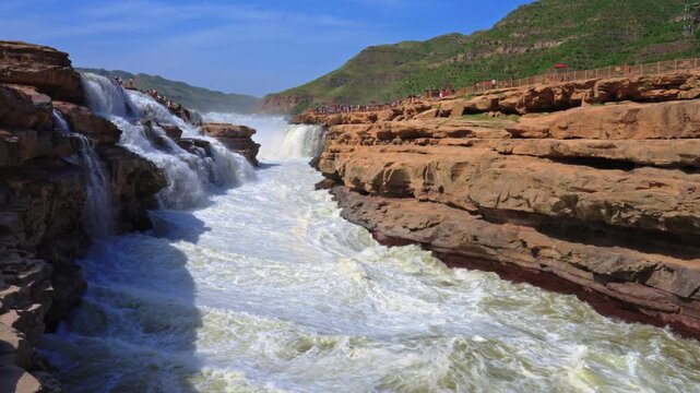 Wide-angle perspective of the Yellow River pouring into the Hukou Waterfall gorge. Tourists observe from the rocky banks as massive cascades create a misty, surging current.