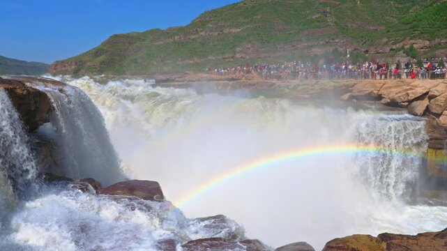 Cinematic wide-angle view of the Hukou Waterfall on the Yellow River. Powerful water cascades into the gorge, creating a prominent double rainbow against a backdrop of rocky cliffs and tourists.