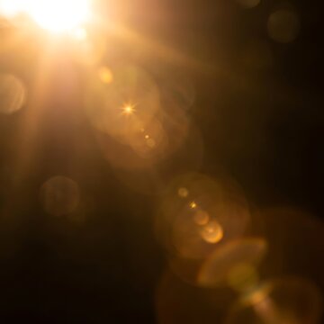 Soft golden sunlight flare with glowing lens flares and warm bokeh effect against a dark, moody background creating a cinematic and dramatic ambiance.