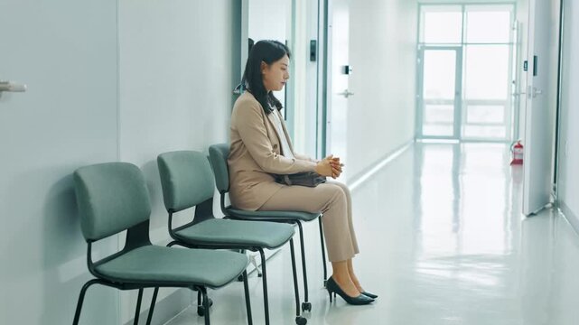 Professional Asian woman waiting patiently in a modern office or hospital hallway, contemplating or anticipating an important meeting, interview, or appointment.