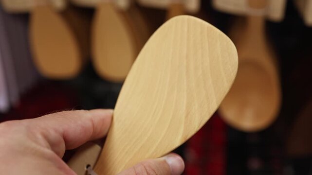 Customer holding a wooden rice paddle spatula at a kitchenware store. Close-up of a hand choosing eco-friendly wood cooking utensils on display in a supermarket department. Stock video footage.