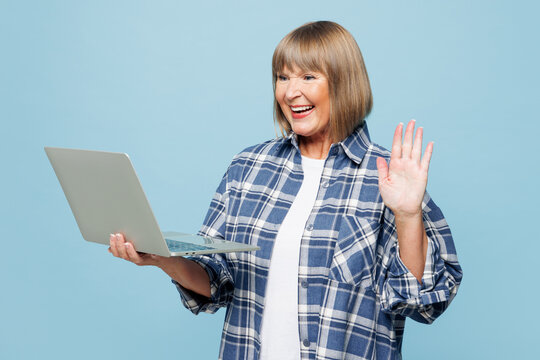 Elderly blonde smart IT woman 50s years old wear checkered shirt casual clothes hold use work on laptop pc computer waving hand talk speak isolated on plain pastel blue background. Lifestyle concept.
