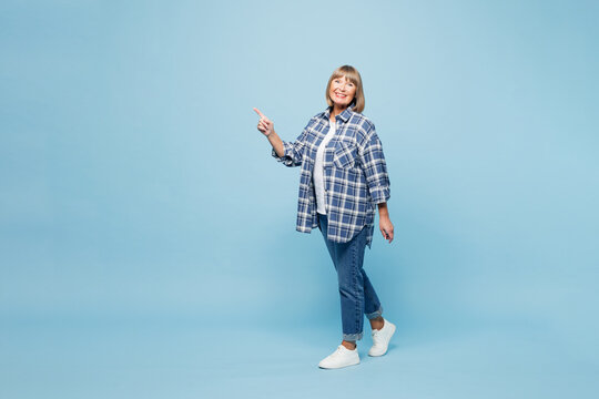 Full body elderly blonde woman 50s years old wears checkered shirt casual clothes point index finger aside on area mock up isolated on plain pastel blue background studio portrait. Lifestyle concept.