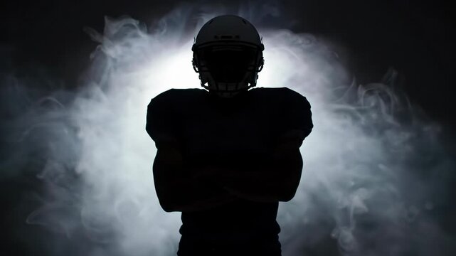 A powerful american football player in full uniform and helmet, arms crossed, stands as a sharp deep black silhouette, backlit in a dark studio with white smoke. Dramatic sports concept.