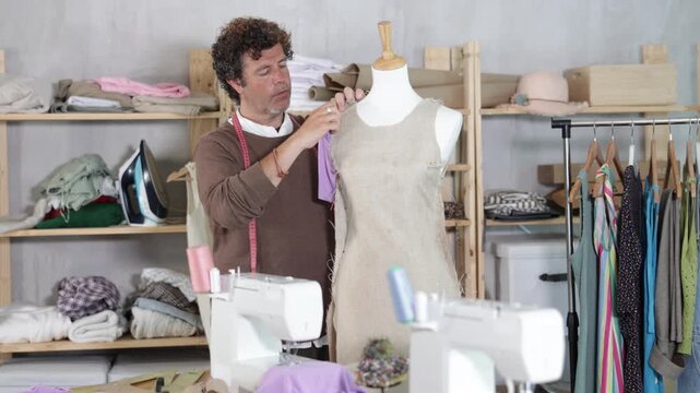 Adult man works as a tailor in a sewing workshop, he stands next to a mannequin and makes edits to a dress sample. Process of tailoring clothes according to an individual sketch and measurements