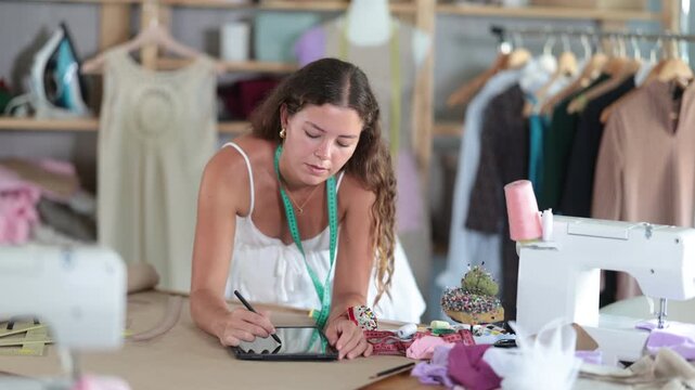 Female dressmaker sits on a table in a workshop and draws a sketch of clothes on an electronic tablet. Designer comes up with a layout once. High quality 4k footage