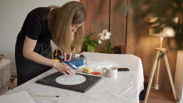 Woman applies white texture paste on black canvas at covered table with art tools nearby. Concept of handmade wall art, DIY decor, and creative process.