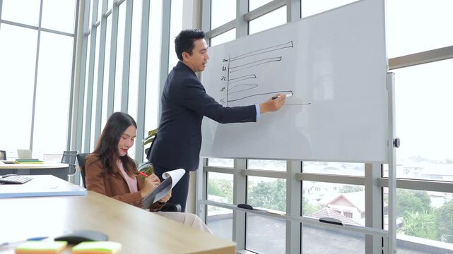Diverse business team collaborating on a new marketing plan. Man explaining ideas on a flipchart while woman taking notes in a bright, glass-wall office.