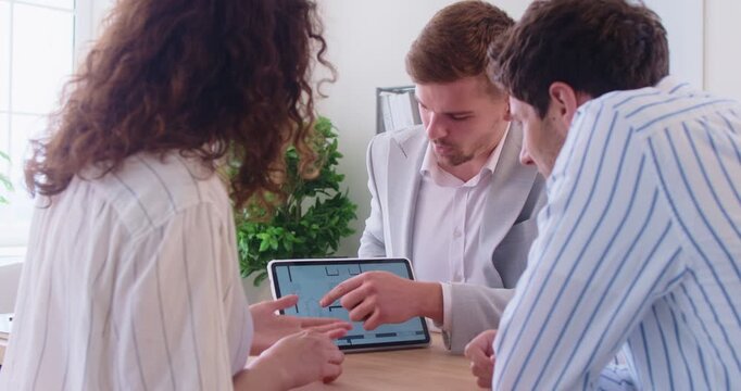 Male realtor showing apartment floor plan on digital tablet to young couple of potential buyers. Man and woman listening and watching layout presentation during real estate consultation in office.