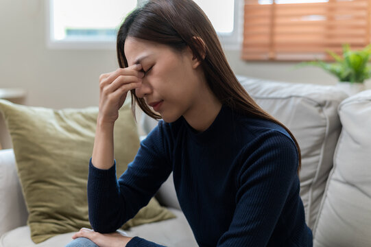 Young Asian woman suffering from headache and dizziness while sitting on sofa at home. Female feeling sick, stressed, exhausted and unwell with migraine, fatigue and health problem concept.