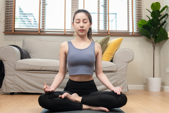 Young Asian woman practicing yoga meditation and mindful breathing at home. Calm female sitting in lotus pose for relaxation, wellness, stress relief and healthy lifestyle concept.