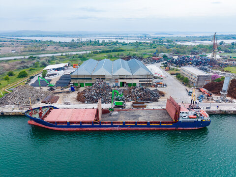 Cargo ship docks at a recycling facility in a coastal area for unloading scrap metal during a cloudy day