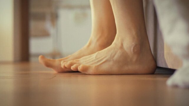 Man slowly rises from bed, stretching his legs before taking a step onto the cool floor barefoot. His morning routine begins with gentle movements, easing into wakefulness on the quiet, domestic scene