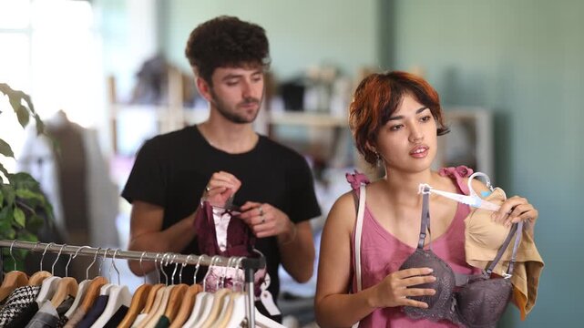 During shared shopping in boutique, young girl examining plain comfortable bra while boyfriend playfully suggesting revealing lace set