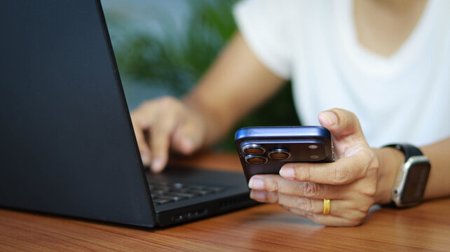Close-up of person using smartphone and laptop at workspace. Concept of remote work, online communication, multitasking, mobile technology, and digital productivity.