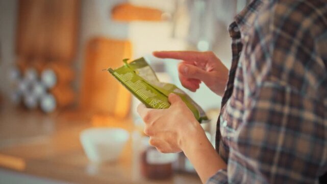 In bright kitchen at midday, girl holds soft plastic packaging, scanning ingredient list. Her careful read highlights healthy lifestyle focus, emphasizing conscious nutrition avoiding harmful foods
