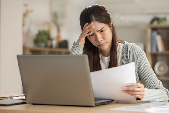 Stressed Asian woman feeling worried about bill and debt. She has financial problem, sitting with laptop at home and holding paper with confused look