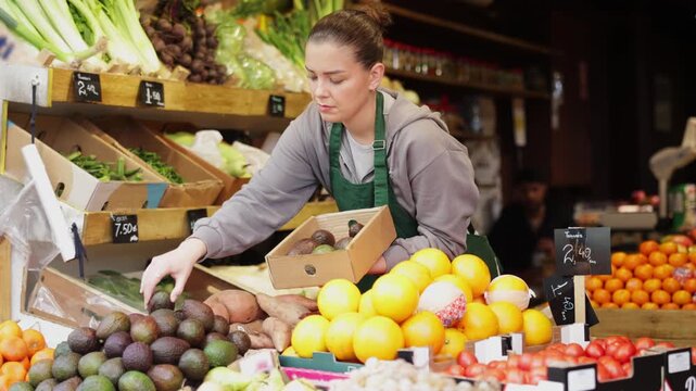 Young woman seller in apron puts fresh avocados on display at vegetable market