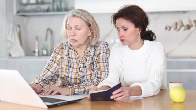 Concentrated old woman showing content of her passport to middle-aged woman sitting next to her and working on laptop in the kitchen