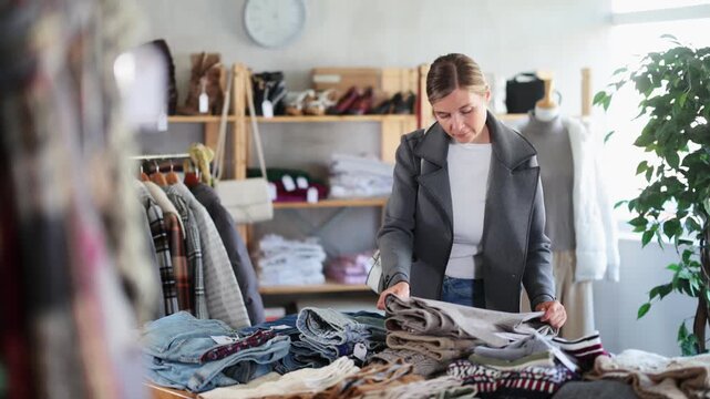  While shopping, woman browsing goods items and choose elect trousers in store, buyer consider some items. Concept of seasonal wardrobe shopping. Client and holiday shopping. 