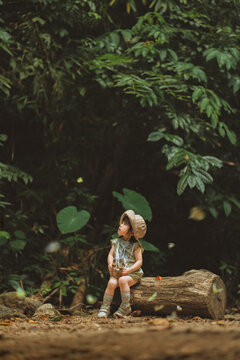 Cute Asian toddler girl holding toy binoculars while sitting on a log, watching flying butterflies at Baan Krang Camp forest. Childhood curiosity, eco tourism, outdoor nature education, adventurous