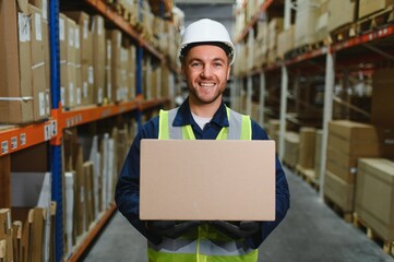 Warehouse worker in blue uniform holding a large cardboard box. Stacks of boxes are visible in the...